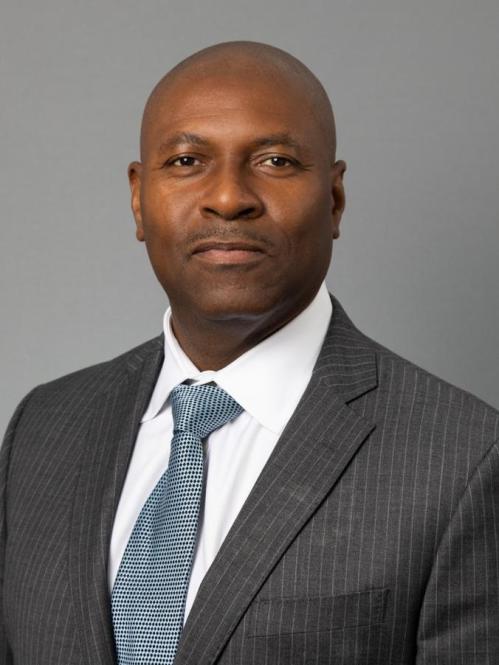 Nayland Smith headshot, in a grey suit and white shirt with a soft blue tie