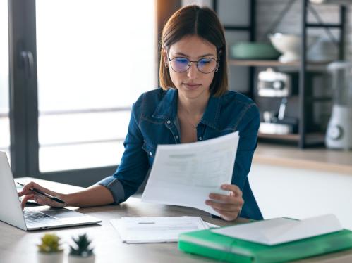 Business woman working with computer while consulting some invoices and documents in the kitchen at home.